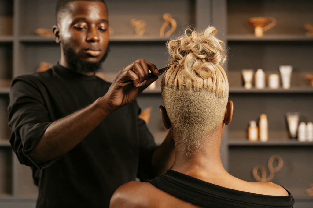 A hair stylist with a short blonde taper styling a client’s curled updo at a salon station.