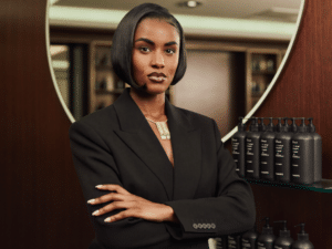 A salon owner in a tailored black blazer standing with arms crossed in front of her product shelf.