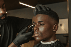 A barber in black gloves using clippers to execute a precision fade on a seated client.