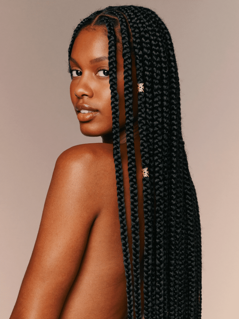 A woman wearing long waist-length box braids with gold cuff accents, photographed in warm studio light.