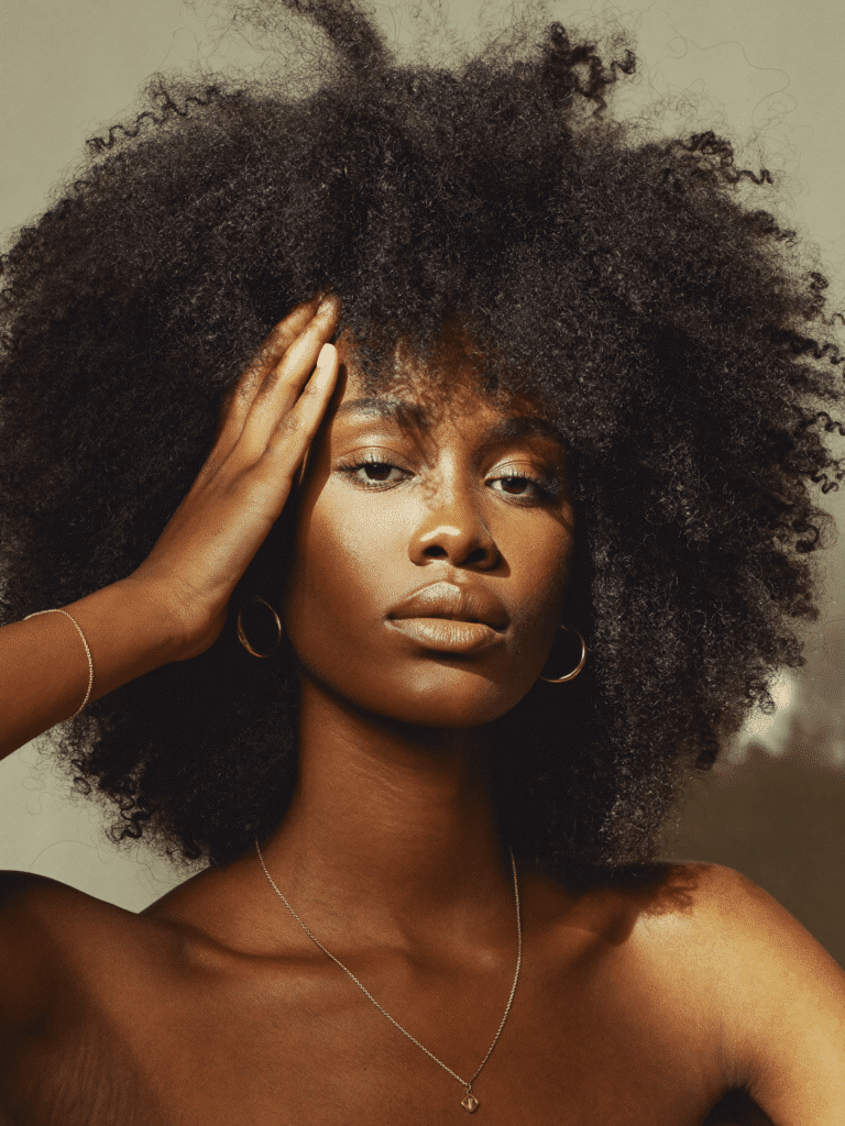 A woman with a full voluminous natural afro photographed against a warm neutral backdrop.
