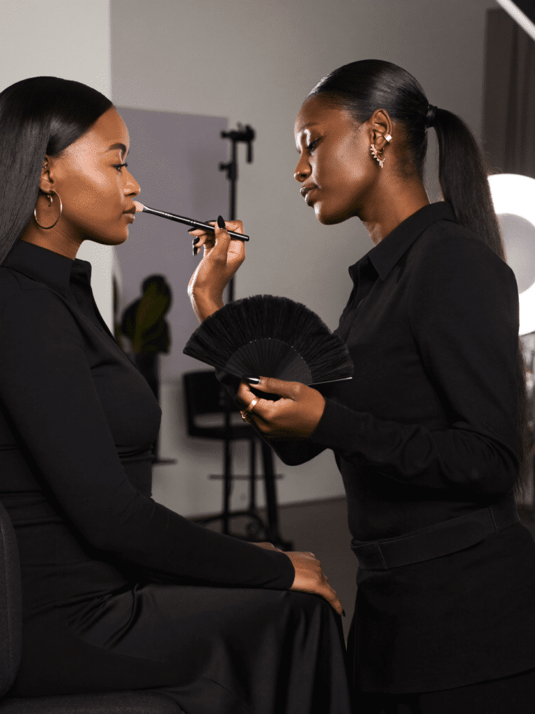 A makeup artist applying highlighter to a seated client’s cheekbone with a fan brush.