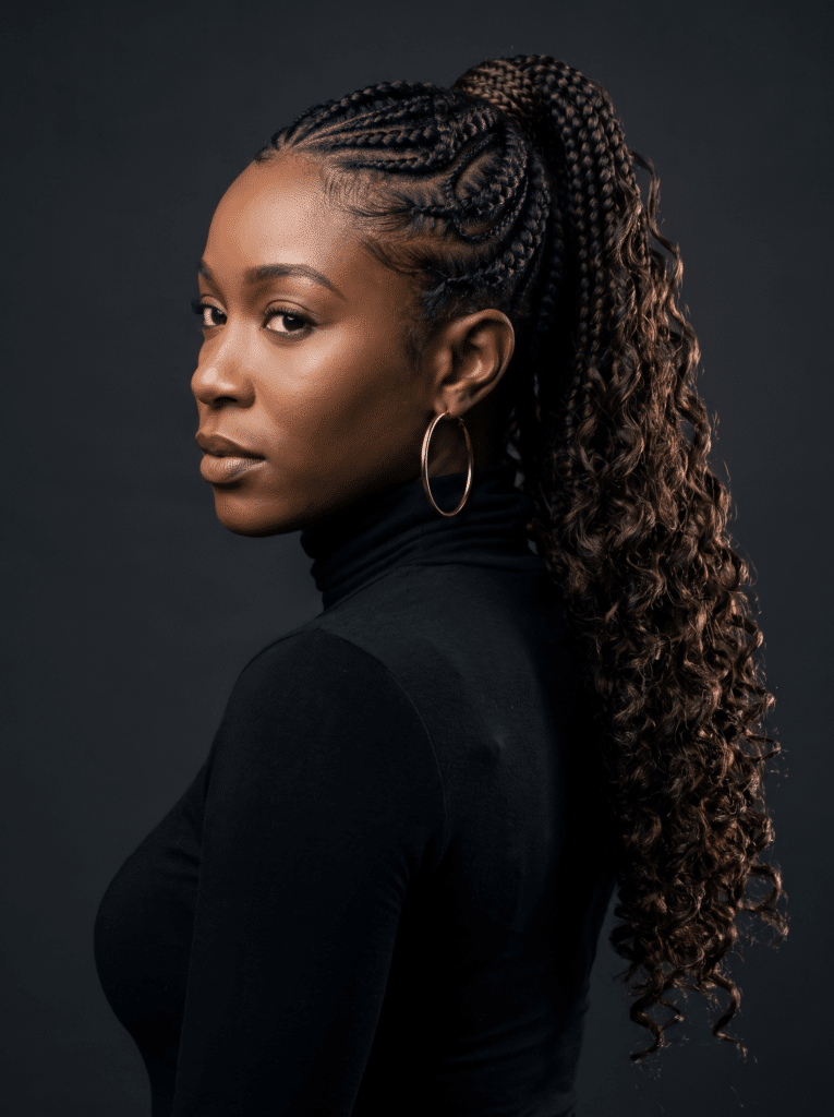 A woman in profile wearing fine cornrow braids flowing into a long curled ponytail, gold hoop earring and black turtleneck.
